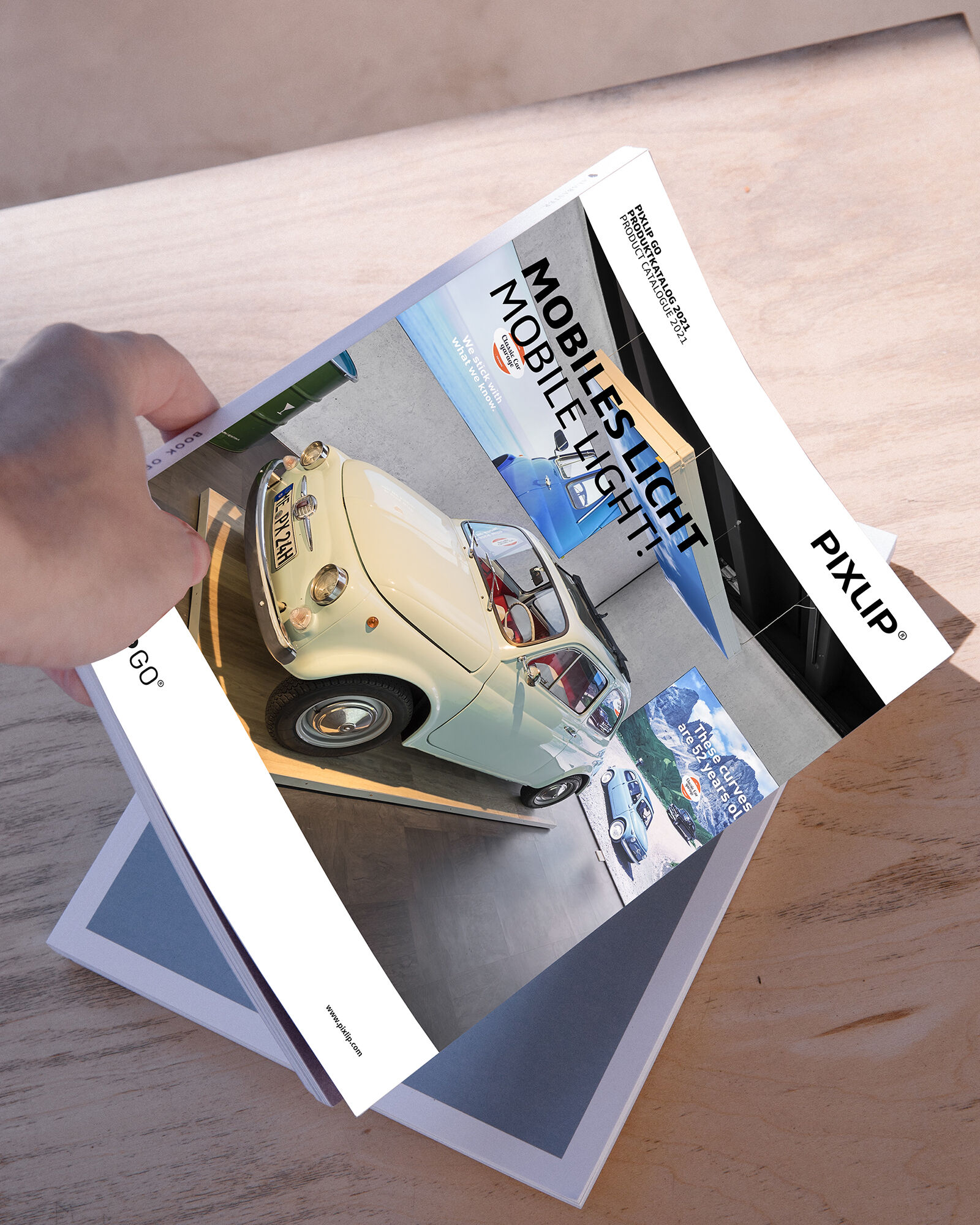 Hand greift nach Katalog von Pixlip mit einem Auto abgebildet, welches mit einem anderen Magazin auf einem Holztisch lag
